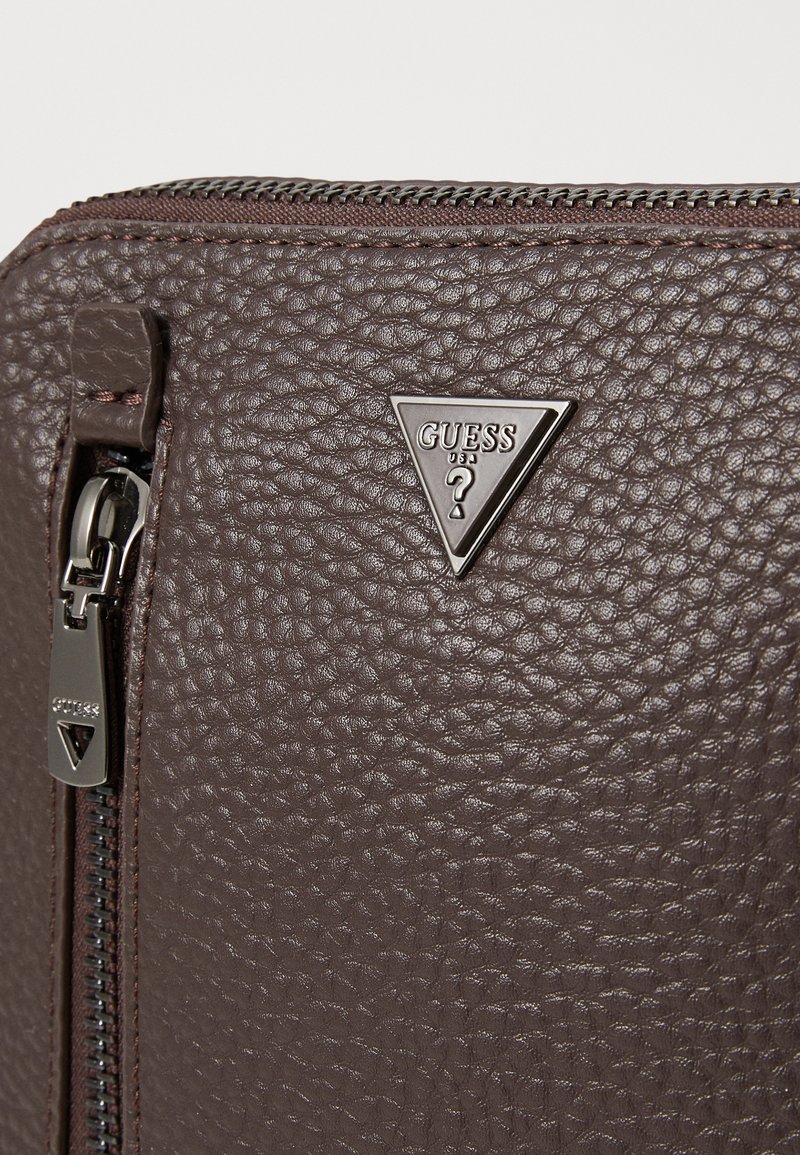 Bolso de cuero marrón texturizado con un cierre de cremallera plateado y una placa de metal en forma de triángulo con el logotipo "GUESS" y un diseño de signo de interrogación.