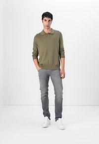 Olivgrünes langärmliges Polohemd mit geripptem Saum, kombiniert mit hellgrauen, figurbetonten Jeans und weißen Sneakers, stehend vor einem weißen Hintergrund.