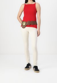 Mujer con una blusa sin mangas roja, un cinturón ancho marrón con grandes medallones decorativos, pantalones ajustados color crudo y zapatillas negras.