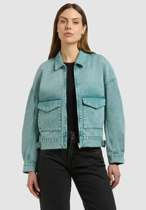 Femeie purtând o geacă scurtă din denim turcoaz cu buzunare mari în față, peste un top negru și blugi negri cu talie înaltă.