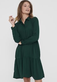 Robe verte foncée à manches longues, à boutons, au design en couches, avec une texture fluide et un col. Tissu lisse avec une coupe décontractée.