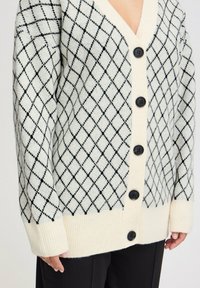 Cardigan crema con un motivo a rombi neri, cinque bottoni neri e polsini a coste. Texture morbida con scollatura a V.