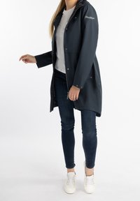 Námořnický nepromokavý kabát s kapucí, zapínáním na zip a knoflíkovými detaily. Kapsy po obou stranách, nošený přes bílé tričko a džíny, doplněný bílými botami.