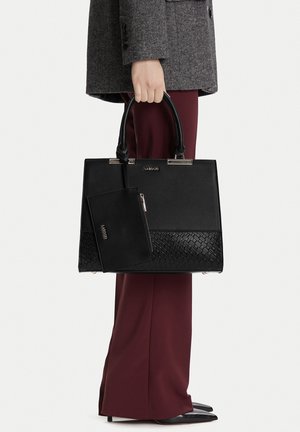 Mano che tiene una borsa nera con texture e pochette abbinata, pantaloni bordeaux, cappotto grigio e scarpe nere a punta su sfondo bianco.