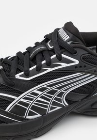 Černá sportovní bota s prodyšným svrškem z síťoviny, bílými detaily a texturovanými tkaničkami. Má výrazný boční proužek a tlumenou podrážku.