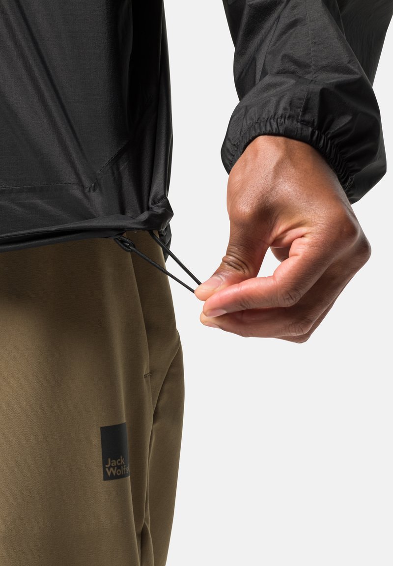 Hand, die den elastischen Kordelzug der schwarzen Jacke anpasst, trägt olivfarbene Jack Wolfskin-Hosen. Nahaufnahme auf Taille und Handgelenk vor weißem Hintergrund.