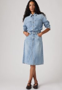 Ανοιχτό μπλε τζιν πουκάμισο με κούμπωμα με κουμπιά και δύο τσέπες στο στήθος, συνδυασμένο με μια αντιστοιχισμένη φούστα σε σχήμα Α. Και τα δύο έχουν λεία υφή και κλασικό σχέδιο.