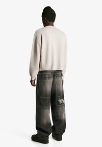 Przesadnie duże szare dżinsy z tylnymi kieszeniami i dekoracyjnym przeszyciem, w połączeniu z jasnoszarym swetrem i czarną czapką beanie.