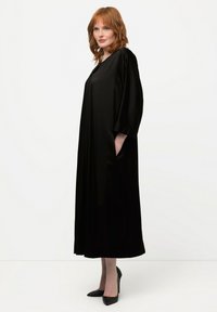 Abito di satin nero con maniche oversize, lunghezza fino al polpaccio e tasche laterali. Indossato con tacchi a punta neri, texture liscia, design minimale.
