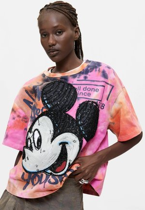 Frau mit geflochtenen Haaren trägt ein pink-orange Batikshirt mit einem großen Pailletten-Mickey-Maus-Gesicht und Textdetails.
