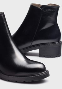 Botines de cuero negro con un acabado brillante, puntera redonda y resistente tacón de bloque negro. Presenta una suela texturizada para un mejor agarre y un discreto logo de la marca.
