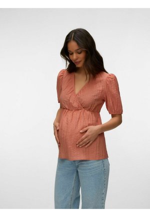 Donna incinta con una camicetta avvolgente color ruggine con trama e jeans azzurri chiari, che tiene delicatamente il pancione e guarda verso il basso.