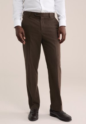 Homme portant un pantalon formel marron foncé, des mocassins noirs et une chemise blanche à manches longues rentrée, debout sur un fond clair.