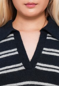 Gros plan sur le bas du visage et le cou d'une personne portant un pull sombre avec des rayures blanches horizontales et un col en V profond.