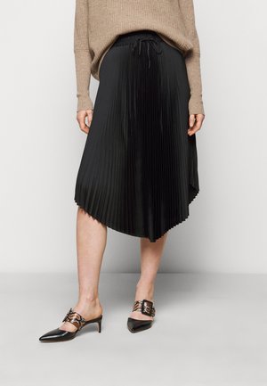 Vrouw met een beige trui, zwarte geplooide asymmetrische midirok en zwarte lage puntige hakken met slangenprint riemen en gespen.