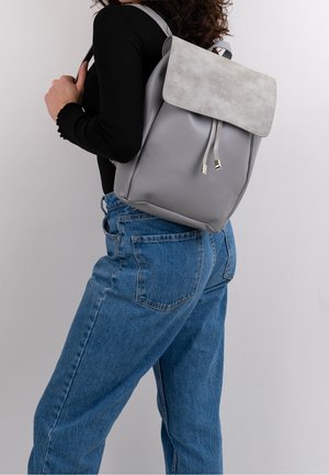 Sac à dos gris avec une surface lisse et un rabat gris clair. Dispose d'une fermeture à cordon et de deux bretelles. Design simple et moderne.