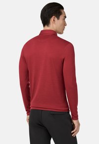 Muž oblečený v upnutém červeném polokošili s dlouhým rukávem a černých kalhotách, viděný zezadu a mírně otočený doprava.