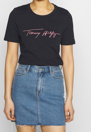 Person wearing a black short-sleeve t-shirt with "Tommy Hilfiger" in red cursive and a high-waisted blue denim skirt with frayed hem.