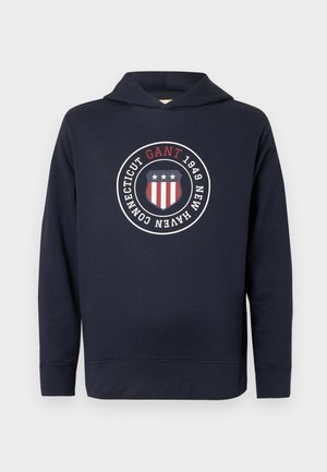 Navyblauwe hoodie van zachte stof. Bevat een rond logo met rode, witte en blauwe accenten, tekst en een trekkoordkap.