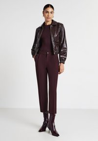 Femme portant un haut côtelé violet foncé, une veste courte brillante violet foncé, un pantalon assorti et des bottines à talons pointus, les mains dans les poches.