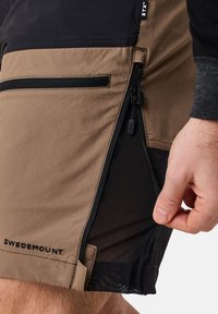 Pantaloni corti khaki e neri con tasche con zip, una texture liscia e un'etichetta intrecciata con "SWEDEMOUNT" sulla cucitura inferiore.