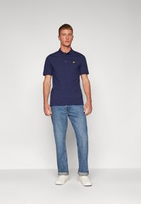 Polo bleu marine avec col, manches courtes et logo jaune. Associé à un jean en denim bleu clair et des baskets blanches.
