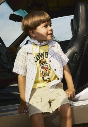 Camisa de cuadros de manga corta en blanco y beige sobre una camiseta gráfica amarilla con un perro de dibujos animados. Pantalones cortos marrones, pañuelo blanco alrededor del cuello.