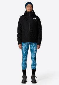 Czarna kurtka puchowa z wysokim kołnierzem, w połączeniu z niebieskimi leggingsami marmurkowymi. Całość dopełniają szare buty trekkingowe oraz niebieska opaska na głowę.