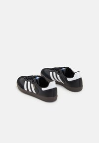 Een paar zwarte Adidas Samba sneakers met witte strepen en bruine zolen, van achteren gezien op een witte achtergrond.