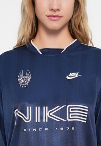 Približeni prikaz osobe koja nosi tamnoplavu Nike sportsku majicu s bijelim logotipom, stiliziranim tekstom "NIKE", i natpisom "Since 1972" ispod.