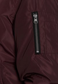 Chaqueta de nailon burdeos con un bolsillo con cremallera lateral. Presenta una textura suave y un hardware de cremallera visible con acentos negros.