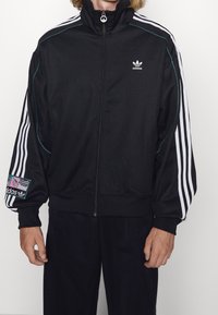 Czarna bluza sportowa rozpinana na suwak z wysokim kołnierzem, białym wzorem z trzema paskami i teksturowaną naszywką na rękawie. Wykonana z gładkiej tkaniny z elastycznymi mankietami.