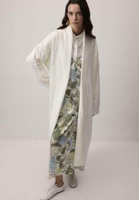 Dlhý biely pletený kardigán cez kvetinové šaty v modrej, zelenej a šedej farbe s bielym golierom, skombinované s bielymi teniskami.