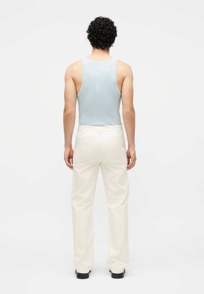 Άνδρας με σγουρά μαλλιά στέκεται με την πλάτη, φορώντας ένα ανοιχτά γαλάζιο τοπ ριμπ, λευκό παντελόνι και μαύρα παπούτσια σε απλό φόντο.