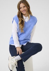 Un gilet en tricot bleu clair avec un col en V, porté sur une chemise blanche à col, assorti avec un jean foncé et des baskets blanches, assis sur une plateforme blanche.