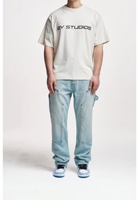 Oversized cream t-shirt with "ZY STUDIOS" print, paired with light blue denim cargo pants featuring pockets, worn with white and blue sneakers.