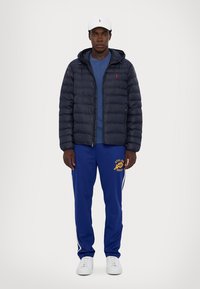 Giacca puffer navy con cappuccio, t-shirt blu a girocollo, pantaloni sportivi blu con logo giallo e sneakers bianche, indossati con un cappellino bianco.