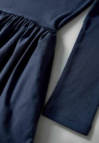 Mörkblå klänning med en figursydd liv med samlad, flytande tyg nedan. Lång ärm med ribbad textur vid manschetten.