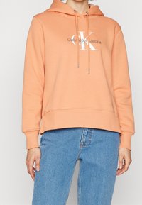 Peach-colored hoodie with a drawstring, cuffed sleeves, and a slightly cropped hem. Features a white "Calvin Klein Jeans" logo on the front.