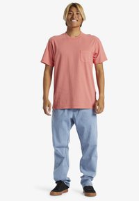 Korálová bavlněná trička s krátkým rukávem a kapsou na hrudi, kombinovaná se světle modrými džíny a černými teniskami s hnědými podrážkami.