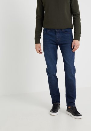 Mann trägt dunkelgrühes Langarmshirt, dunkelblaue Jeans und schwarze Sneaker, steht vor einfarbig hellem Hintergrund.