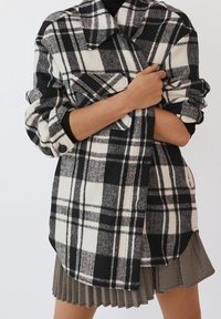 Camisa de franela a cuadros en blanco y negro con cuello, mangas enrolladas y detalles de botones. Lleva puesta sobre una blusa negra de canalé y una falda con estampado.