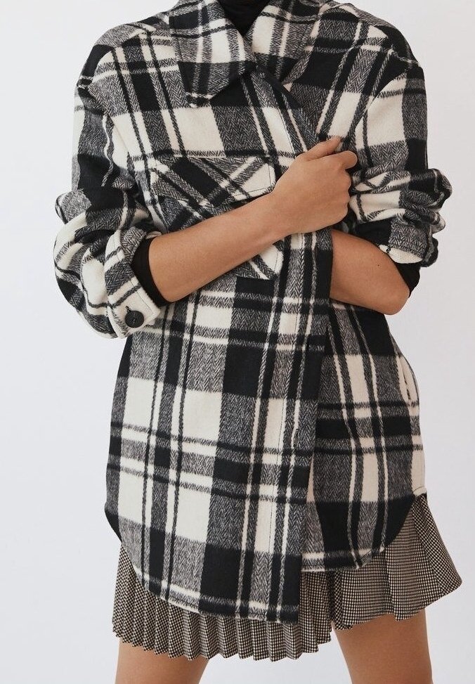 Camisa de franela a cuadros en blanco y negro con cuello, mangas enrolladas y detalles de botones. Lleva puesta sobre una blusa negra de canalé y una falda con estampado.