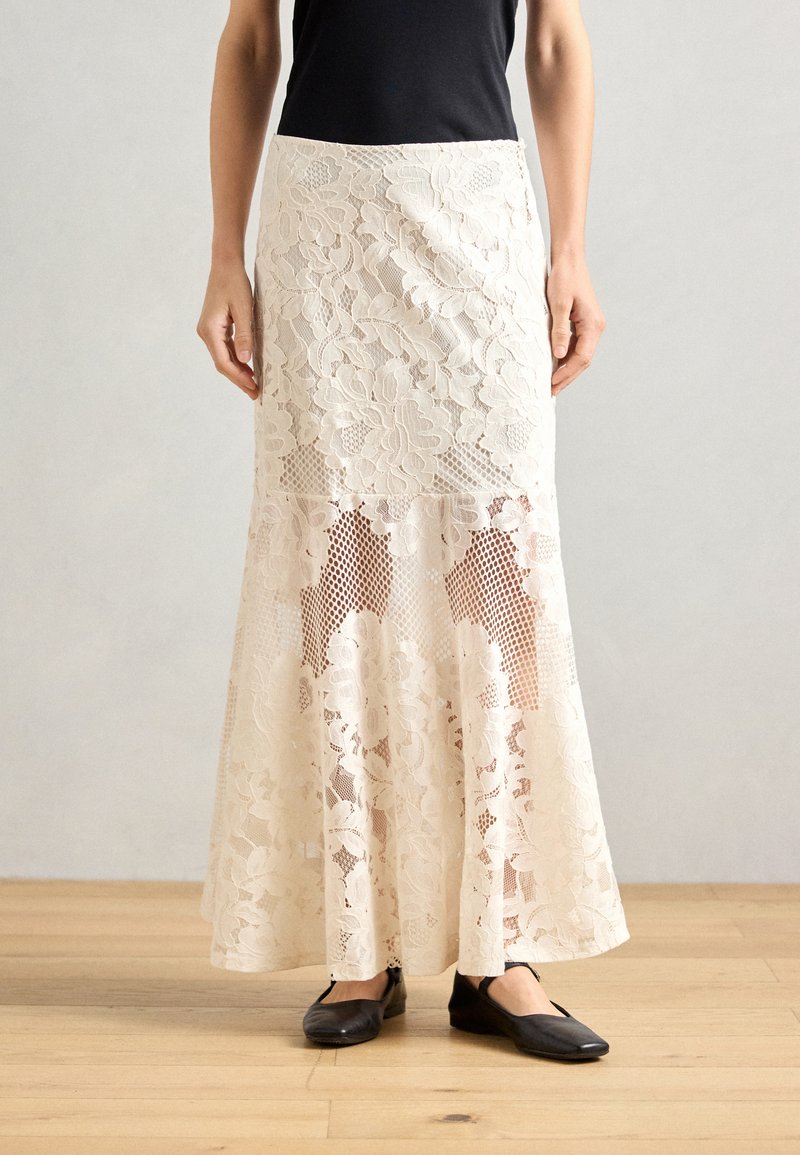 Lace maxi skirt in ivory with floral patterns and sheer panels, featuring a fitted waist and flared hem. Worn with black flats.