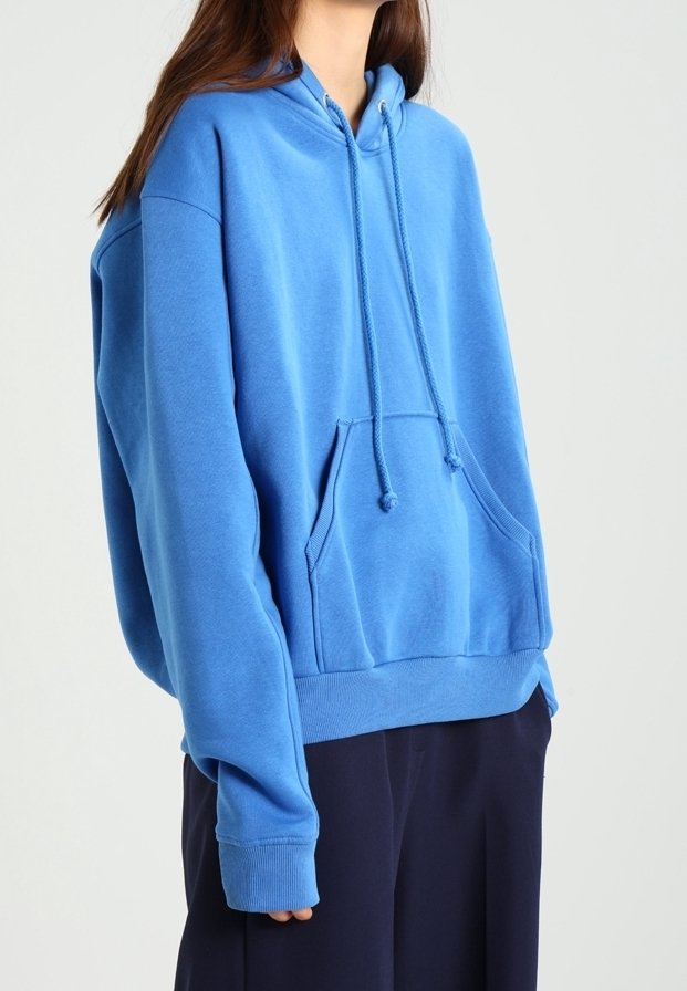Femme portant un sweat à capuche bleu clair oversize avec poche avant et cordons, associé à un pantalon ample foncé sur un fond uni.
