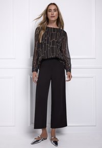 Czarna, przezroczysta bluzka z długim rękawem i geometrycznym wzorem; zestawiona z czarnymi, szerokimi spodniami z wysokim stanem oraz błyszczącymi, spiczastymi klapkami.