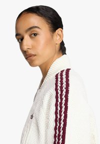 Cardigan bianco con trama e collo a coste, con dettagli a strisce bordeaux e bianche sulle maniche e piccolo logo vicino al petto.