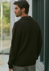 Pull en maille marron foncé avec une coupe décontractée, un large col et des poignets côtelés. Porté avec un jean gris clair, mettant en valeur un tissu texturé.