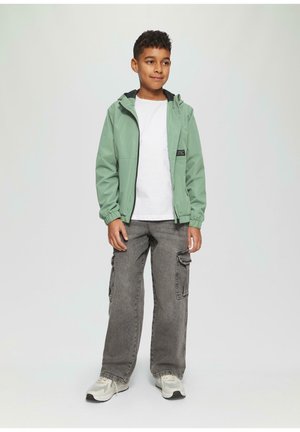 Grüne, leichte Jacke mit Kapuze, Reißverschluss vorne und elastischen Bündchen, kombiniert mit einem weißen T-Shirt und grauen Cargo-Hosen. Neutrale Sneaker.
