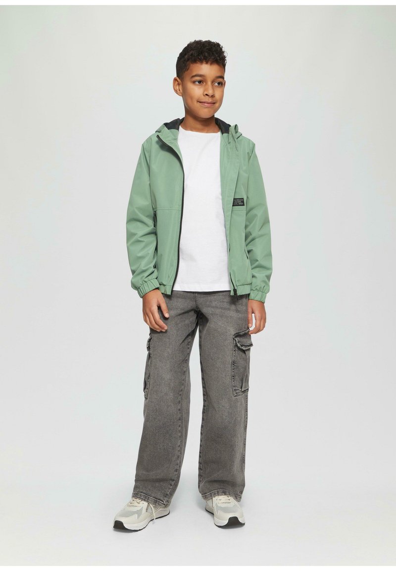 Grüne, leichte Jacke mit Kapuze, Reißverschluss vorne und elastischen Bündchen, kombiniert mit einem weißen T-Shirt und grauen Cargo-Hosen. Neutrale Sneaker.